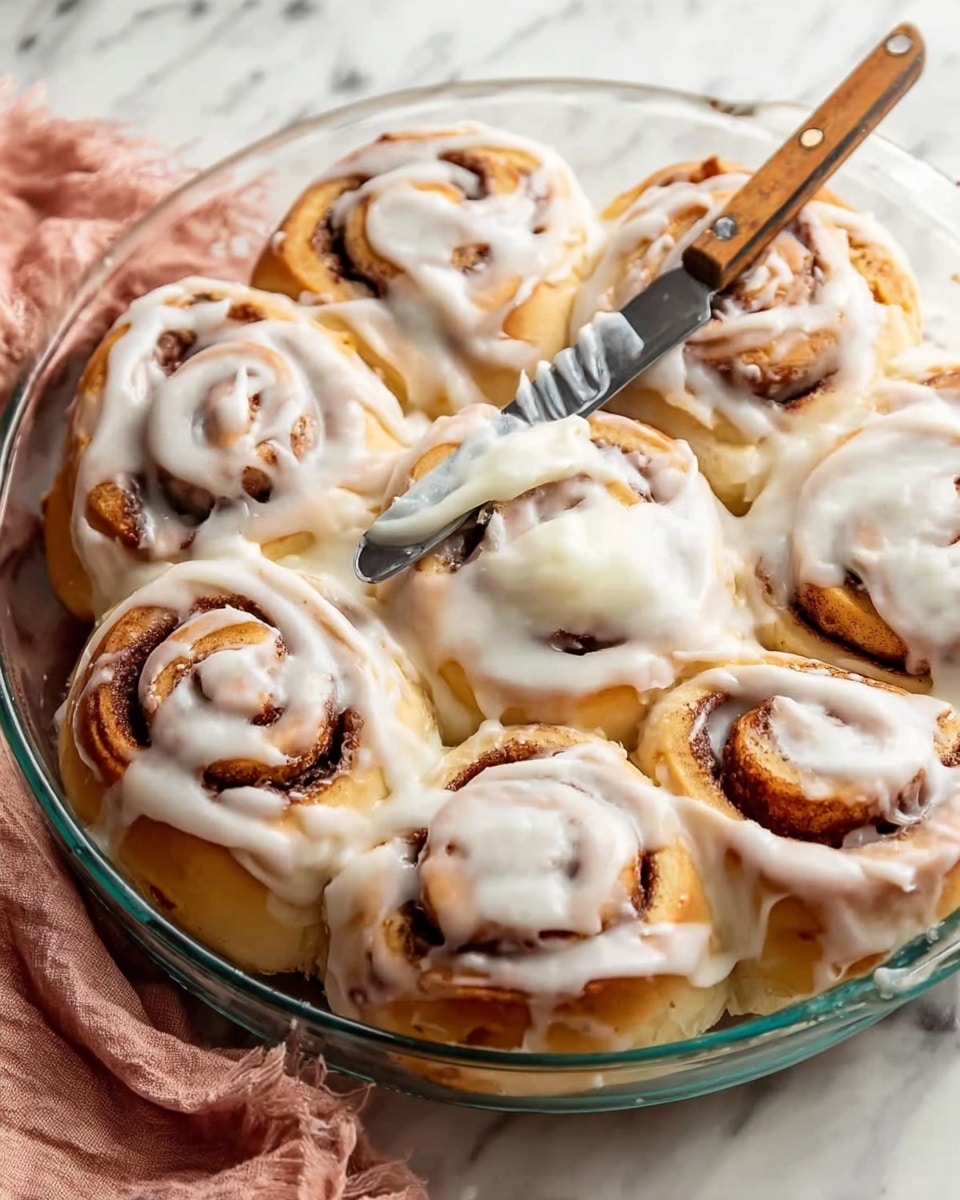 A round glass dish filled with nine cinnamon rolls baked golden brown, each topped thickly with creamy white icing that looks smooth and soft. The cinnamon rolls show distinct swirls of brown cinnamon filling visible through the light tan dough. A silver knife with a wooden handle rests on top, spreading more white icing on one of the cinnamon rolls. The dish is placed on a white marbled surface with a soft pink towel partially seen at the bottom left corner. photo taken with an iphone --ar 4:5 --v 7