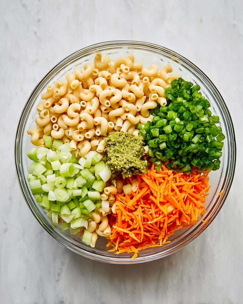 A clear glass bowl on a white marbled surface shows five separate layers of ingredients, arranged side by side like sections of a circle. One layer is light tan elbow macaroni with a curved shape and smooth texture. Next to it, a small dollop of green-greenish paste sits on top of the pasta. Adjacent to this is a layer of chopped bright green onions with a fresh and crisp look. Another section holds finely diced pale green celery pieces that look firm. The last section contains a pile of bright orange shredded carrots with a thin, stringy texture. The bowl is viewed from above, showing all layers clearly. Photo taken with an iphone --ar 4:5 --v 7