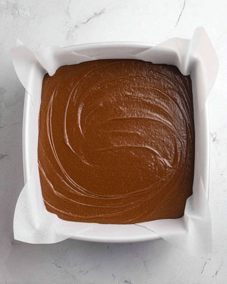 A single thick layer of smooth, rich brown batter evenly spread inside a white baking pan lined with white parchment paper. The batter surface shows soft, long swipes and slightly shiny texture, filling the pan almost to the edges. The pan is set against a white marbled surface, creating a clean and simple look. photo taken with an iphone --ar 4:5 --v 7