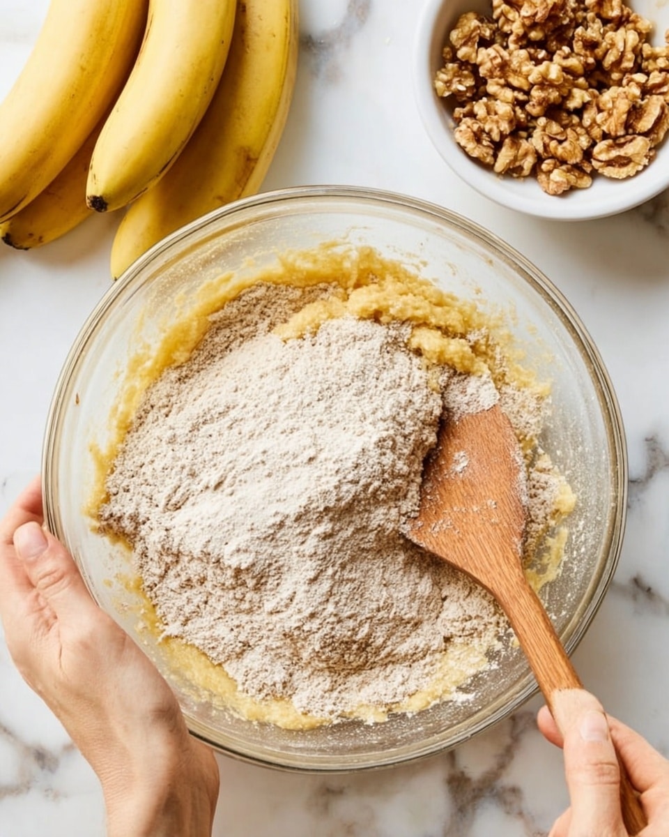 A clear glass bowl holds a thick mixture with two main layers: the bottom layer is a yellowish mashed banana texture, partly covered by the top layer of light brown flour mix with some flour still dry and powdery. A woman's hand on the right side holds a wooden spoon stirring the batter, while another woman's hand on the left side steadies the bowl. In the background on a white marbled surface, there are two ripe yellow bananas on the top left and a white bowl filled with chopped walnuts on the top right. The photo taken with an iphone --ar 4:5 --v 7
