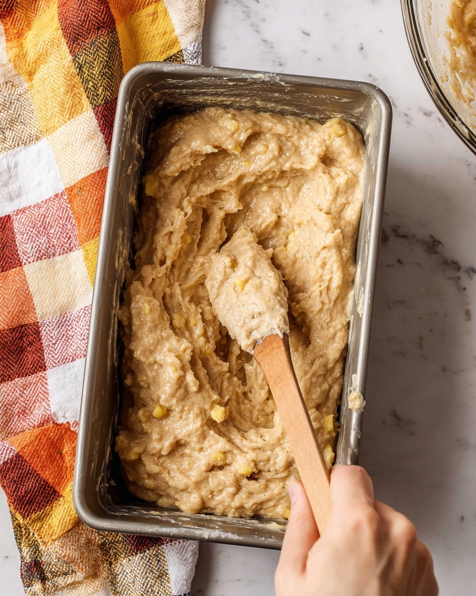 The image shows a metal rectangular baking pan filled with thick, lumpy light brown batter that has some yellow chunks mixed in. A wooden spoon with batter coating its round end is resting inside the pan, held by a woman's hand at the bottom right corner. The pan sits on a white marbled surface next to a colorful checkered cloth in shades of orange, yellow, white, and black. A glass mixing bowl with remaining batter parts is partially visible on the right edge. photo taken with an iphone --ar 4:5 --v 7