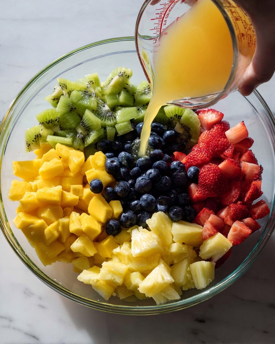 A clear glass bowl filled with five sections of fresh, chopped fruits on a white marbled surface: bright green kiwi pieces at the top, yellow mango chunks on the left, dark blue blueberries below the mango, pale yellow pineapple pieces at the bottom right, and red strawberries to the right of the kiwi. A woman's hand is pouring light orange juice from a glass measuring cup into the bowl, adding a fresh touch to the colorful fruit mix. The lighting is natural, making the colors vivid and fresh. photo taken with an iphone --ar 4:5 --v 7