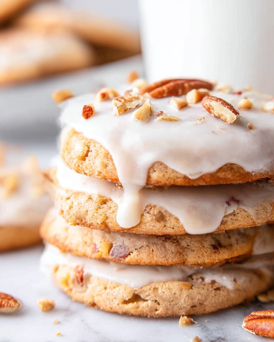 A close-up of a stack of four thick cookies on a white marbled surface, each cookie light brown with visible bits of nuts inside, showing a rough texture. The top cookie is covered with smooth white icing that slightly drips over the edges and is sprinkled with small pieces of chopped nuts. In the background, there is a blurred white container holding more cookies. photo taken with an iphone --ar 4:5 --v 7