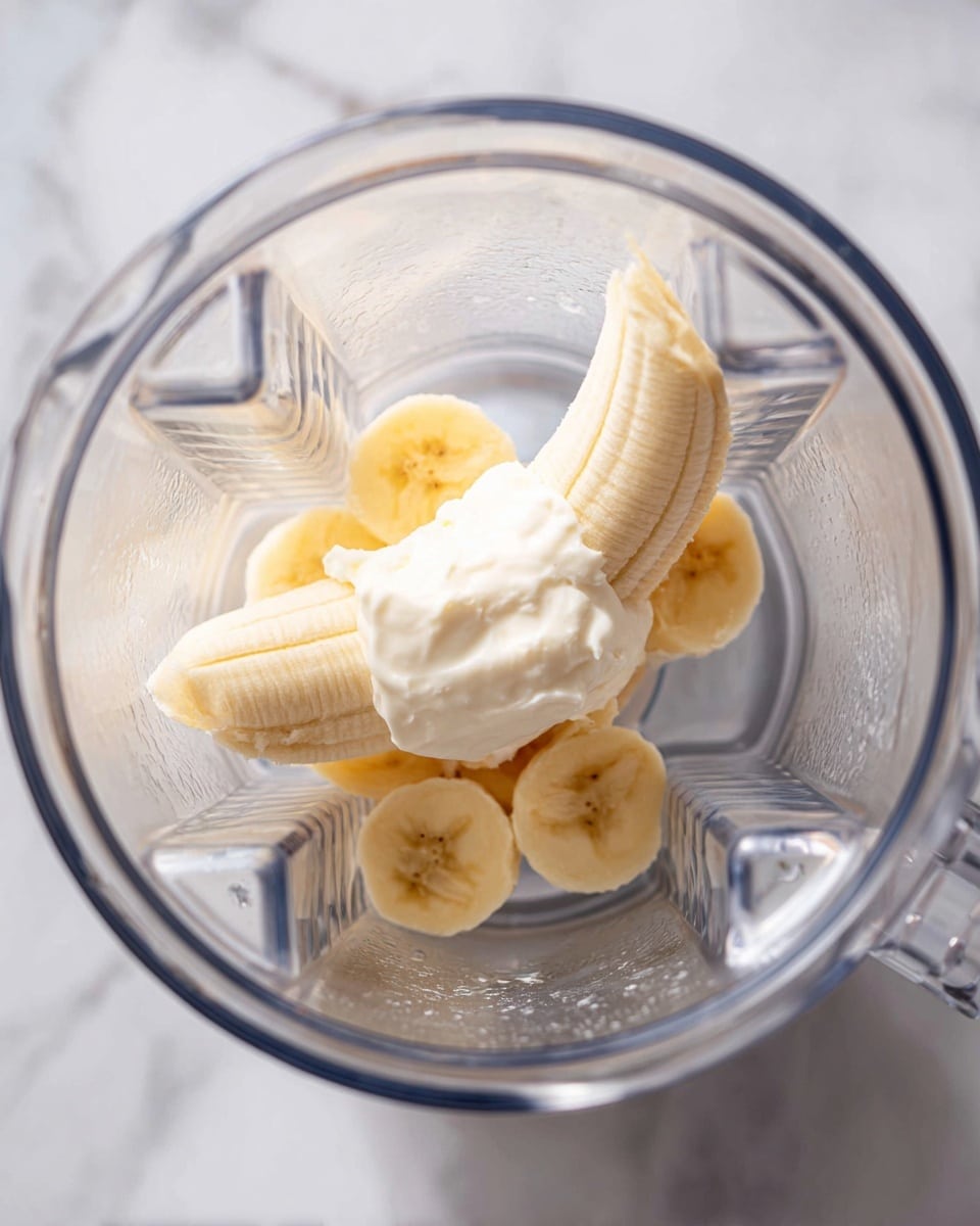 Inside a clear blender container, there is one peeled banana with a small dollop of thick, white cream or yogurt on top, both placed centrally. The banana's pale yellow and slightly textured surface contrasts with the smooth, creamy white topping. The blender container’s inside shows angular ridges and a somewhat shiny, reflective surface. The background is a white marbled texture. photo taken with an iphone --ar 4:5 --v 7
