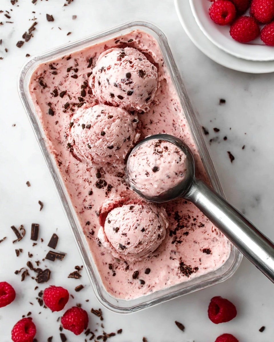 The image shows a clear rectangular container filled with creamy pink ice cream that has small dark chocolate pieces mixed throughout. Three rounded scoops of the ice cream sit on top, with a metal scoop lifting one scoop from the container. Around the container, fresh red raspberries and scattered dark chocolate shavings lay on a white marbled surface. In the top right corner, a white plate with a few raspberries is partially visible. Photo taken with an iphone --ar 4:5 --v 7
