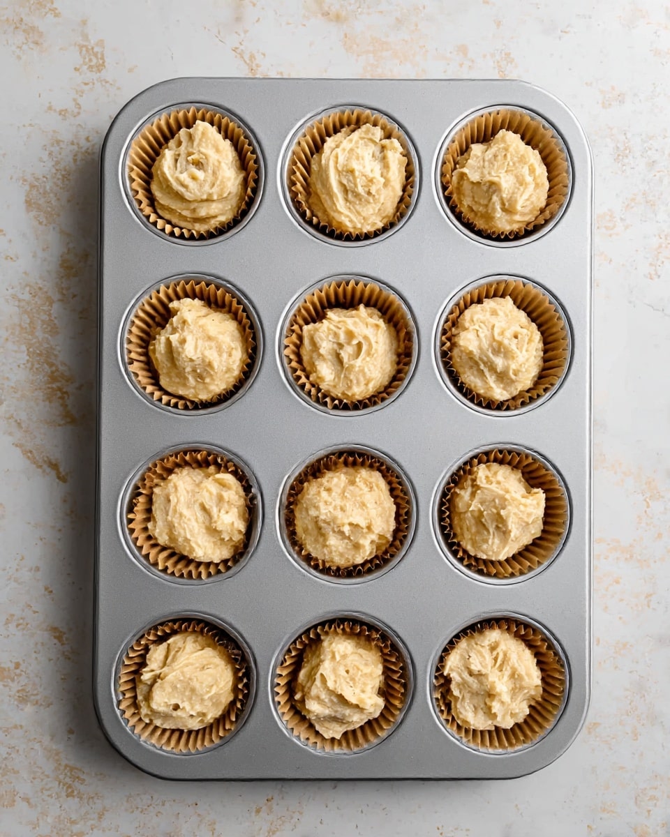 A metal muffin tray with twelve round cups, each lined with a brown paper baking cup filled with light beige, slightly textured batter that looks creamy and thick. The muffin tray is placed on a white marbled surface with soft, uneven gray and beige spots. The batter in each paper cup is slightly rough and swirled on top, showing small lumps and a few air pockets, filling each cup about three-quarters full. The photo is taken from above, showing the full tray and all the batter cups evenly spaced. Photo taken with an iphone --ar 4:5 --v 7