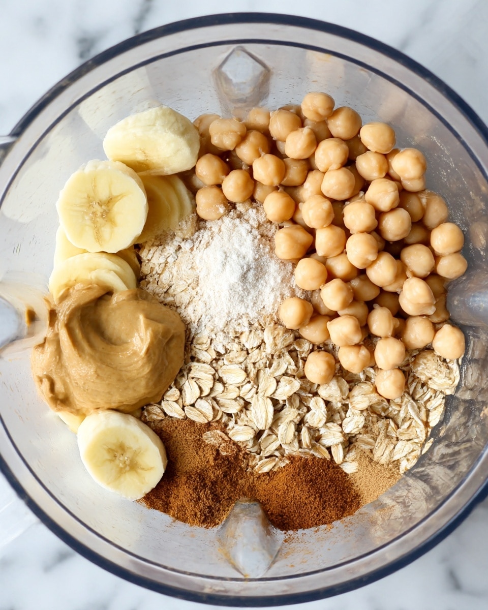 A clear food processor bowl filled with several layers of ingredients: a base layer of light tan rolled oats on the right side, next to beige chickpeas piled in the upper left section, a small pile of white powder on chickpeas, three pieces of peeled yellow banana placed around the bowl, a dollop of creamy light brown peanut butter on the left, and several piles of brown cinnamon powder sprinkled near the peanut butter and oats, all sitting on a white marbled surface photo taken with an iphone --ar 4:5 --v 7