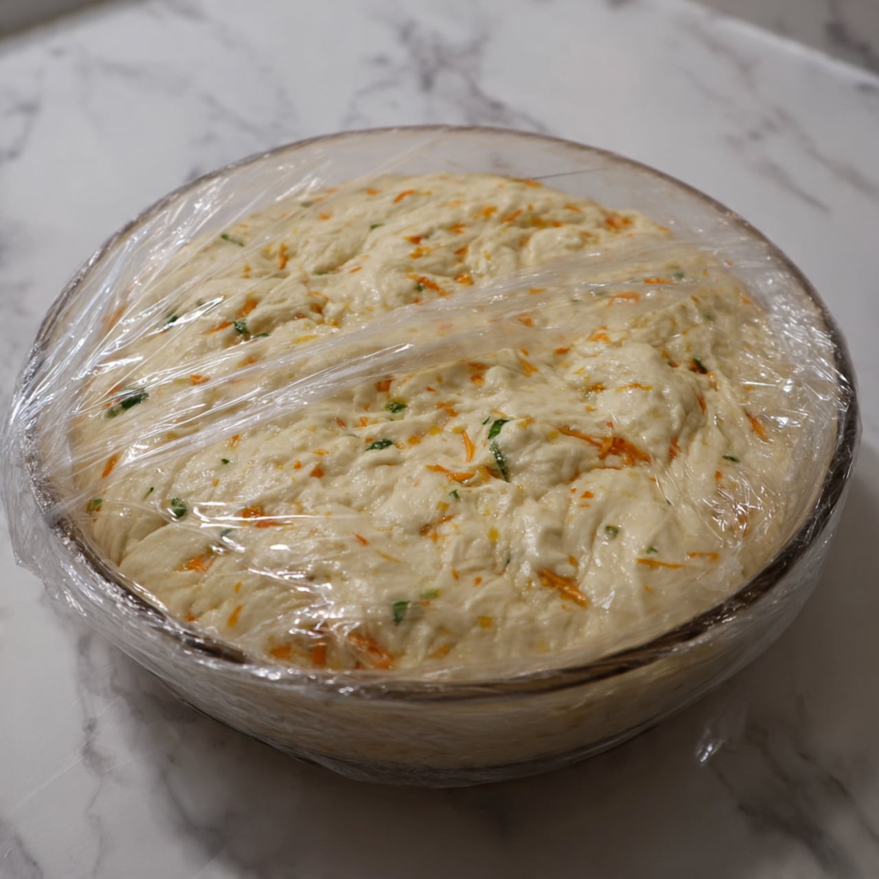 The image shows a clear glass bowl filled with dough that has risen and stretched, with a thick, airy texture full of small holes and a light beige color mixed with orange specks and some green bits. The dough covers the bowl's bottom and sides unevenly, with visible pull marks that look sticky and soft. The bowl is sitting on a white marbled surface, and the dough is partially covered with transparent plastic wrap that clings to the edges of the bowl. The background also has a soft white marbled texture. photo taken with an iphone --ar 4:5 --v 7