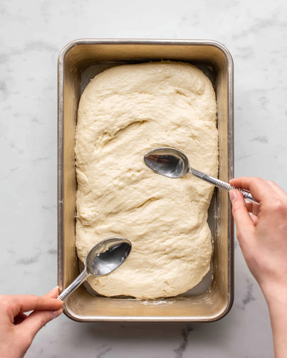 A light beige dough is spread inside a rectangular baking pan with a slightly shiny surface. The dough is wide and roughly oval, with a smooth but slightly uneven texture. Two spoons are pressing and shaping the dough at the top right and bottom left corners, with a woman's hands holding each spoon. The baking pan sits on a white marbled surface, giving a clean and bright look. photo taken with an iphone --ar 4:5 --v 7
