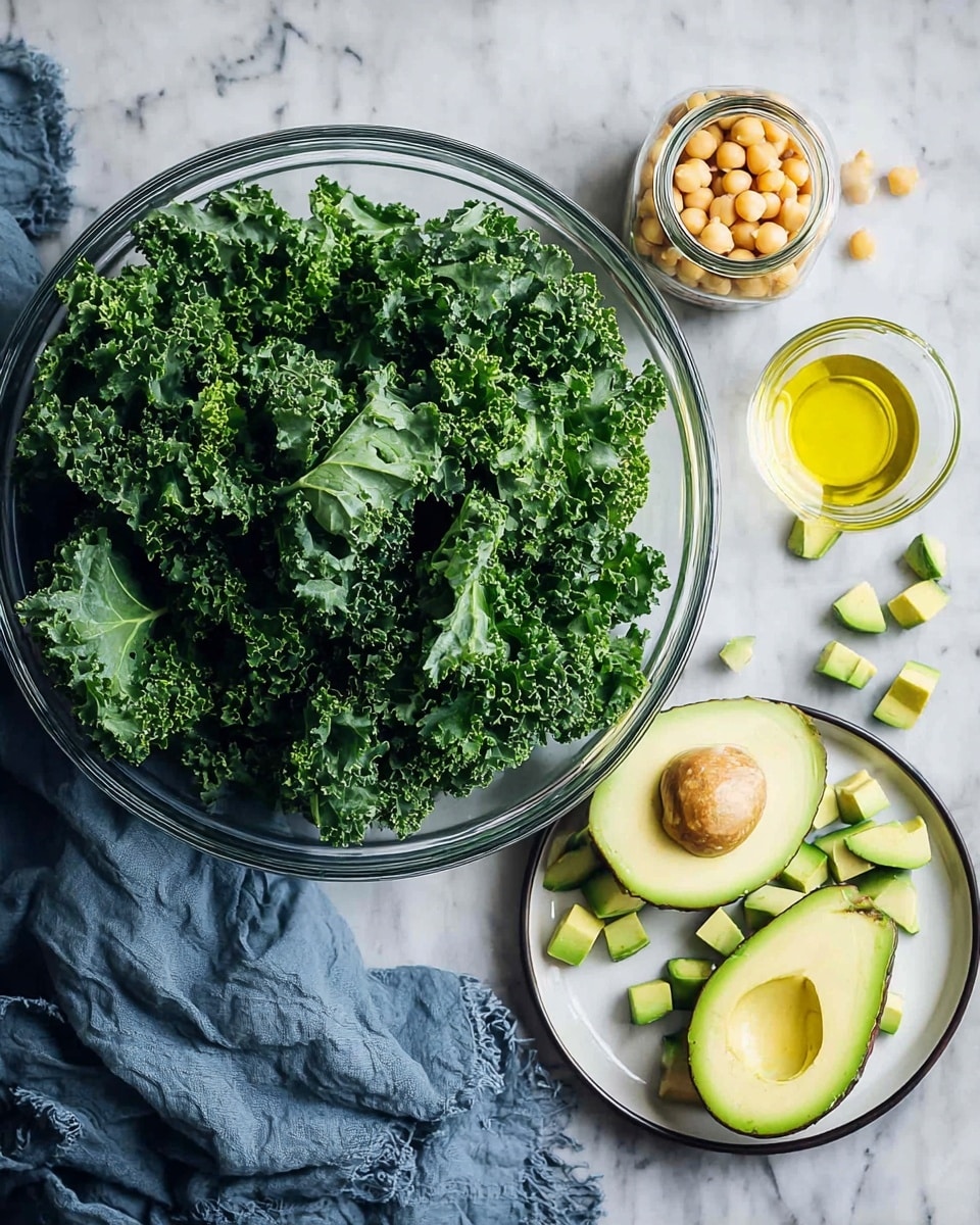 A large clear glass bowl filled with fresh curly dark green kale leaves sits on a white marbled surface. To the right, a small open glass jar contains pale beige chickpeas, and below it, a tiny glass jar holds light yellow oil. Next to the jars, there is a white plate holding half a green avocado with the pit in place, and several small diced avocado cubes scattered beside it. A soft, crumpled blue cloth rests to the right of the plate, adding a cozy touch. Photo taken with an iphone --ar 4:5 --v 7
